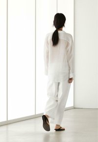 Person with long dark hair in a ponytail walks away wearing a sheer white shirt, loose white pants, and black sandals in a bright, minimalist room.