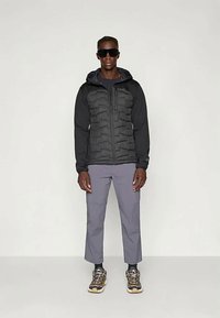 Čierna vypchávaná bunda s kapucňou a kontrastným dizajnom rukávov, kombinovaná so sivými nohavicami a turistickými topánkami. Slnečné okuliare dopĺňajú celý outfit.