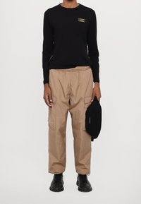 Černé tričko s dlouhým rukávem a zlatým logem, béžové cargo kalhoty s elastickým pasem, černé tenisky a černá taška přes rameno.