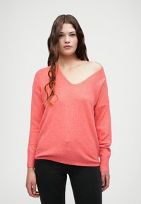 Suéter grande de color rosa coral con un escote en V profundo, puños acanalados y una textura suave, combinado con pantalones negros ajustados.