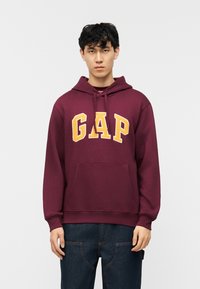 Νέος άνδρας φορώντας μπλούζα με κουκούλα μπορντό με τη λέξη "GAP" σε μεγάλα κίτρινα γράμματα, συνδυασμένη με σκούρο μπλε τζιν, στέκεται μπροστά σε απλό λευκό φόντο.