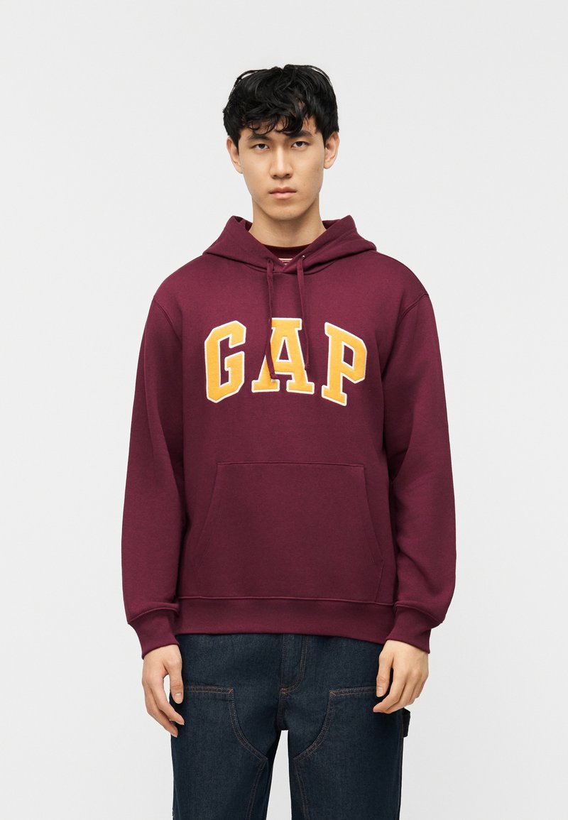 Νέος άνδρας φορώντας μπλούζα με κουκούλα μπορντό με τη λέξη "GAP" σε μεγάλα κίτρινα γράμματα, συνδυασμένη με σκούρο μπλε τζιν, στέκεται μπροστά σε απλό λευκό φόντο.