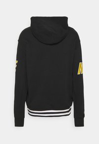 Svart huvtröja med en vit och svart randig fåll, med stor gul grafisk text på vänster ärm. Mjuk tygkvalitet, avslappnad passform.