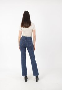 Jeans w kolorze niebieskim, typu flare, z wysokim stanem, z dwoma tylnymi kieszeniami i standardowym paskiem. Noszone z dopasowaną jasną bluzką i czarnymi szpilkami.