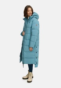 Lys blå pufferjakke med hette, med glidelås og trykknapper, quiltet tekstur og sidelommer. Båret over mørke jeans og beige støvler.