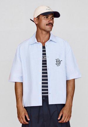 Mężczyzna w jasnoniebieskiej koszuli z krótkim rękawem zapinanej na guziki, na czarno-białej paskowanej koszulce i beżowej czapce z daszkiem z logo.