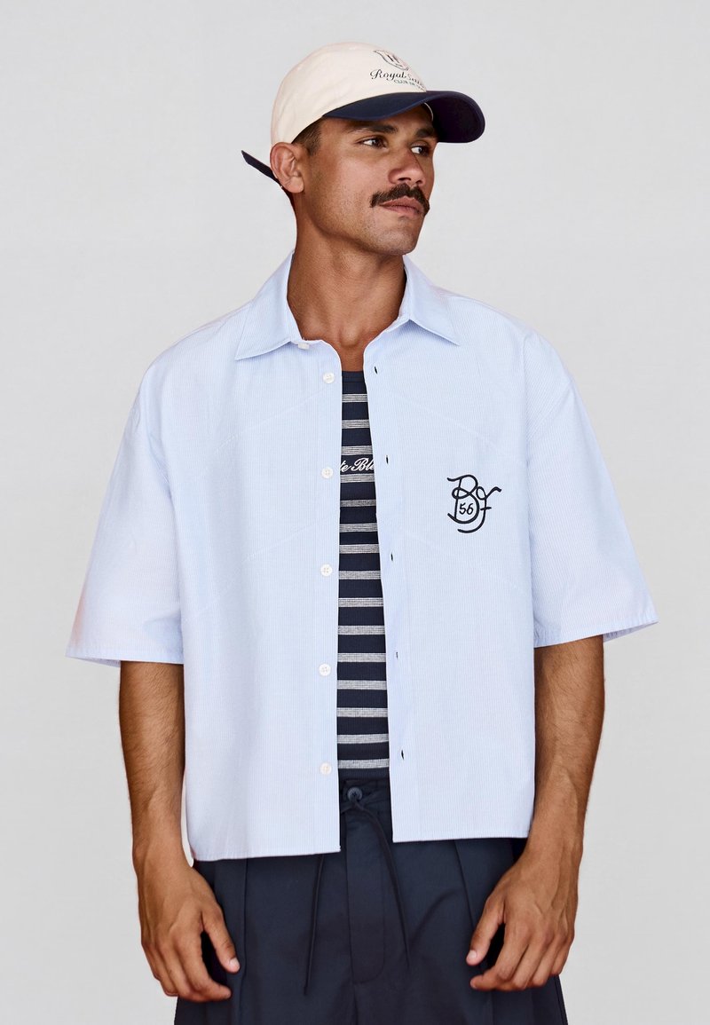 Homme portant une chemise à manches courtes bleu clair boutonnée par-dessus un t-shirt rayé noir et blanc et une casquette de baseball beige avec un logo.