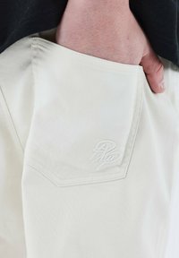 Pantalones beiges con una tela texturizada, con un bolsillo frontal que presenta un logo bordado en blanco. Una mano visible en el bolsillo añade contexto.