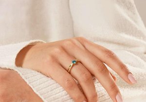 Gold ring featuring a central blue gemstone, with a textured band and small accent stones. Supported by a hand resting on a white fabric.