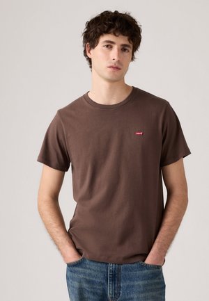 Jonge man met krullend haar, gekleed in een bruine Levi's t-shirt en blauwe jeans, staande met handen in de zakken tegen een effen achtergrond.