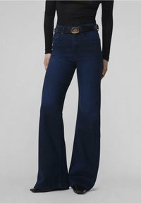 Jeans acampanados de cintura alta en denim azul oscuro. Presenta un cinturón negro con una hebilla dorada circular. Textura suave, ajustados en la cintura.