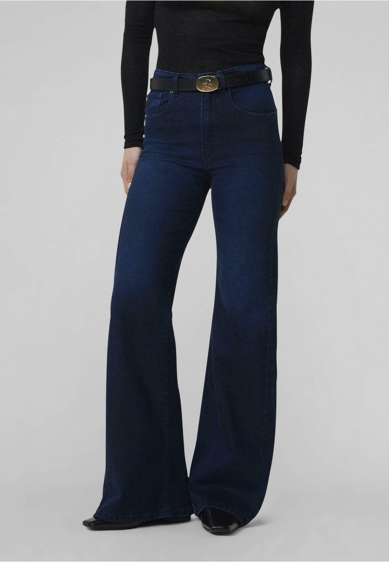 Jeans acampanados de cintura alta en denim azul oscuro. Presenta un cinturón negro con una hebilla dorada circular. Textura suave, ajustados en la cintura.