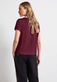 Kvinde med langt blondt hår iført en kortæ兽t maroon skjorte og sorte bukser, stående med hænderne i lommerne, vendt væk.