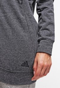 Primer plano de una mano al lado de un sudadera de Adidas de color gris oscuro con un logo visible cerca del dobladillo en el lado izquierdo.