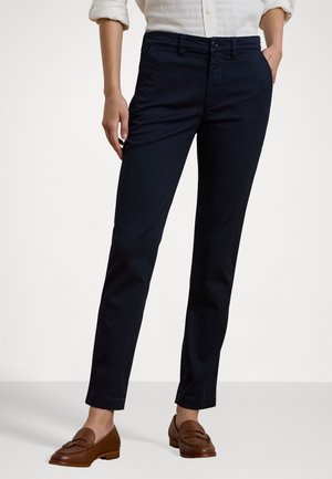 Vrouw die zwarte, slanke broek, witte opgerolde mouwen van een overhemd en bruine leren loafers draagt, staand met één hand in de zak.