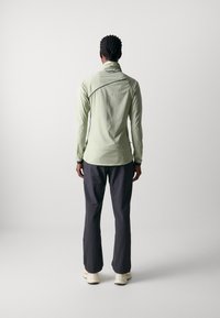 Haut long vert clair avec un col montant et des accents noirs, associé à un pantalon gris foncé et des chaussures de couleur claire. Texture lisse.
