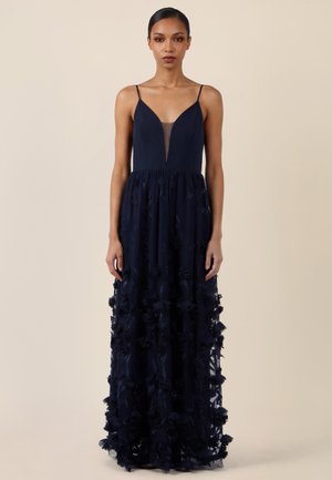 Vrouw staat en draagt een mouwloze marineblauwe jurk met een diepe V-hals en 3D bloemenapplicaties op de rok tegen een effen beige achtergrond.