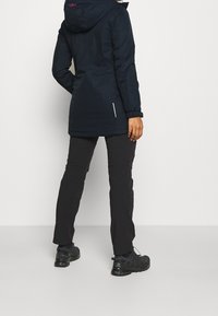Veste longue isolante bleu marine avec capuche à cordon, poignets élastiques et deux poches latérales. Associée à un pantalon noir et des chaussures de sport noires.