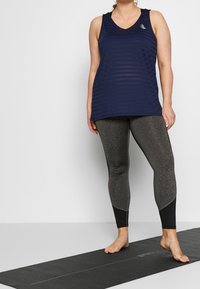 Marineblaues Sporttanktop mit strukturiertem Design, kombiniert mit dunkelgrauen Leggings mit schwarzen Akzenten. Das Modell steht auf einer Yogamatte.