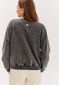 Šedá oversized mikina s ribovanými manžetami a lemem; na zadní straně má malou barevnou karikaturu. Měkká, texturovaná látka.