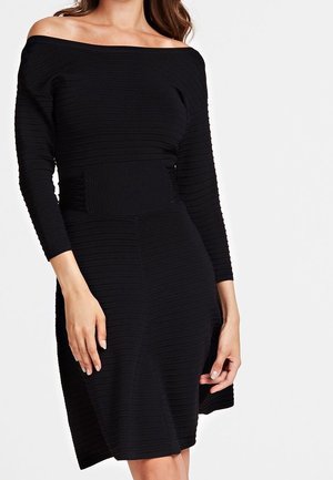 Kobieta ubrana w czarną, prążkowaną sukienkę odsłaniającą ramiona, z długimi rękawami i dopasowanym paskiem w talii, stojąca na jednolitym białym tle.