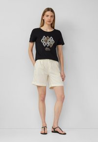 Frau steht vor einer schlichten Wand, trägt ein schwarzes Grafik-T-Shirt, beigen Shorts mit umgeschlagenem Saum und schwarze Sandalen, die Hände in den Taschen.