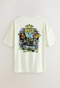 Vaalean kerma väriin painettu puuvillapaita, jossa on värikäs graafinen suunnittelu, jossa on hahmoja, auto ja teksti "OUT KAST" ja "BOB" rohkeassa typografiassa.