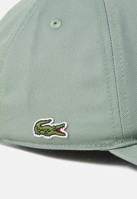 Grønn baseballcaps i teksturert stoff, med et fremtredende brodert krokodillelogo i grønn med røde detaljer.