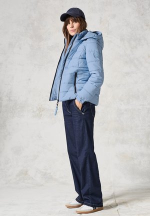 Giacca a vento blu chiaro con cappuccio, chiusura a zip e zip neri; abbinata a jeans di denim scuri e sneakers bianche. Berretto in testa.