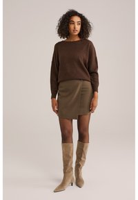 Bruine gebreide trui, olijfgroene wikkelrok en beige kniehoge haklaarzen. Heeft zachte texturen en een getailleerde silhouet.