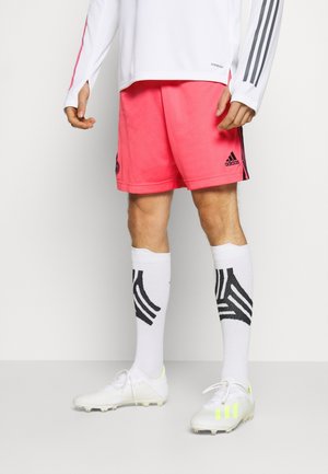 Osoba ubrana w biały sportowy top z długim rękawem, jasnoróżowe szorty Adidas, białe podkolanówki z czarnymi wzorami oraz białe korki z neonowymi detalami.