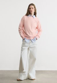 Νέα γυναίκα που φοράει ροζ πουλόβερ πάνω από μπλε ριγέ πουκάμισο με φαρδιά λευκή παντελόνα και ταμπά παπούτσια, στέκεται με τα χέρια στις τσέπες.