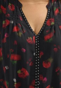 Svart blus med röda och rosa blommönster, V-ringning, plisseringar och en rad silvernitar framtill. Slät, lättviktig tyg.