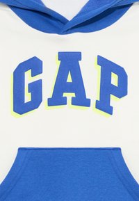 Un hoodie blanc avec un logo "GAP" bleu et jaune néon sur le devant, une poche bleue en bas et une capuche bleue.
