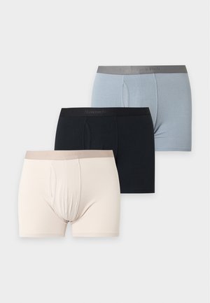 Homme noir mince debout pieds nus portant un boxer noir, regardant de côté sur un fond uni gris clair.
