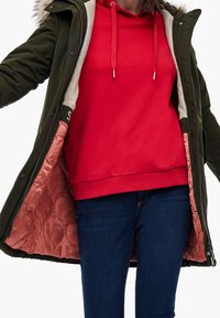 Chaqueta parka acolchada en verde oliva con capucha de piel sintética. Forrada con tela de punto en tono rosa empolvado. Combinada con una sudadera roja y jeans azules.