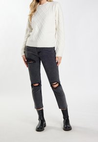 Femme portant un pull en maille blanc, un jean noir déchiré et des bottines noires, debout devant un fond blanc uni.