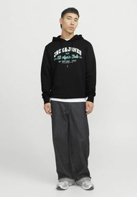 Sudadera negra con gráfico en blanco y verde azulado de "Jack & Jones Surfer's Club", combinada con jeans oscuros holgados y zapatillas grises.