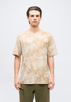 Młody mężczyzna z zaczesanymi do tyłu włosami, ubrany w beżową koszulkę tie-dye i oliwkowo-zielone spodnie typu cargo, stoi na tle jednolitego tła.