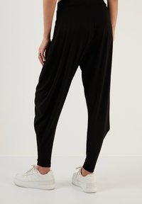 Zwarte, loszittende harem pants met een taps toelopend silhouet, zachte textuur en een strakke tailleband, gedragen met witte sneakers.
