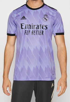 Mężczyzna ubrany w fioletową koszulkę piłkarską Realu Madryt z czarnym kołnierzem i rękawami oraz czarne spodenki Adidas ze złotymi paskami.
