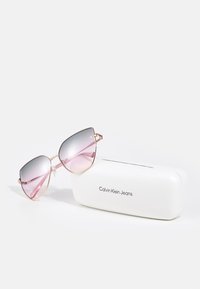 Gafas de sol tintadas en rosa con montura dorada reposando sobre un estuche blanco rectangular con la marca Calvin Klein Jeans sobre un fondo liso y claro.