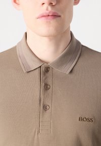 Polo bege feito de tecido texturizado, com colarinho clássico, pala de três botões e logotipo "BOSS" bordado em castanho.