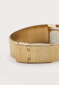 Χρυσός μπρασελές ρολογιού Calvin Klein με κούμπωμα και ραβδωτή υφή σε λευκό φόντο.