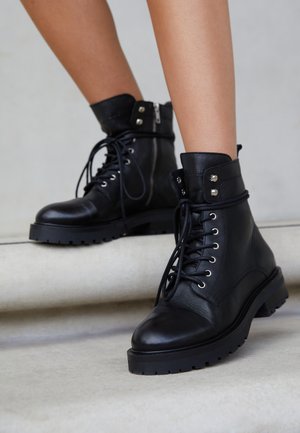 Bottines en cuir noir à lacets avec fermeture éclair latérale, portées sur jambes nues, debout sur une marche en béton clair.