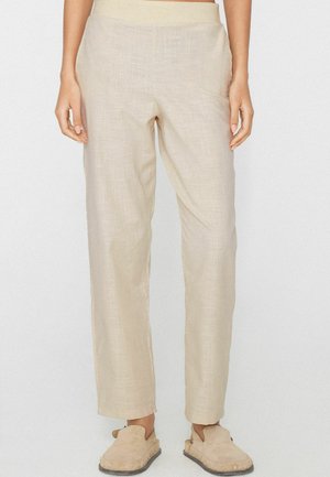 Beżowe lniane spodnie o prostych nogawkach noszone z jasnobeżowymi butami wsuwanymi na osobie stojącej na jednolitym tle.