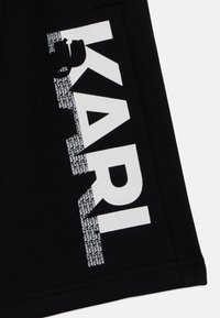 Zwarte stof met gedurfde witte letters die "KARI" spellen, met daaronder kleinere tekst in een herhaald patroon, wat een grafisch ontwerp effect creëert.