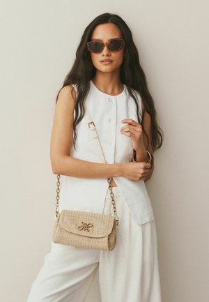Beżowa pleciona torebka na łańcuszku, z klapką i ozdobnymi elementami metalowymi. Modelka ubrana w biały top bez rękawów i okulary przeciwsłoneczne.