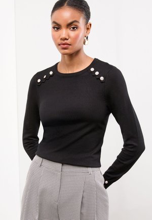 Vrouw draagt een zwarte top met lange mouwen met decoratieve zilveren knopen op de schouders en manchetten, gecombineerd met grijze geruite hoge taille broek.