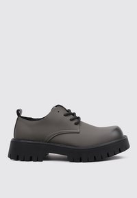 Zapato derby de cuero sintético gris con cordones negros, suela negra gruesa y detalles texturizados en los laterales. Diseño de punta redondeada.